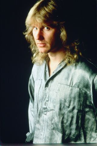 A man with long hair poses in a silver shirt against a dark background.