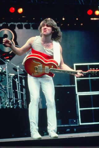 Musician performing energetically on stage, wearing a tank top and white pants while playing guitar.