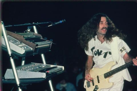 Musician plays bass guitar on stage beside synthesizers during a live performance.