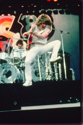 Musician energetically performs with a guitar, leaping on stage while a drummer plays behind.
