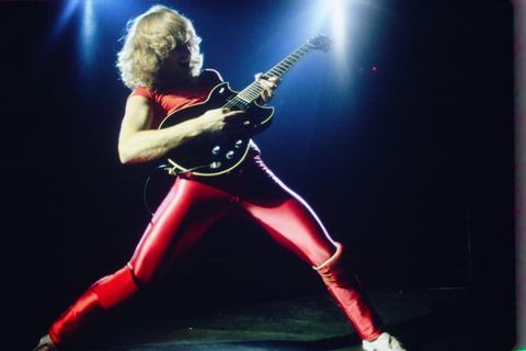 Guitarist in red outfit passionately performs on stage under bright lights.